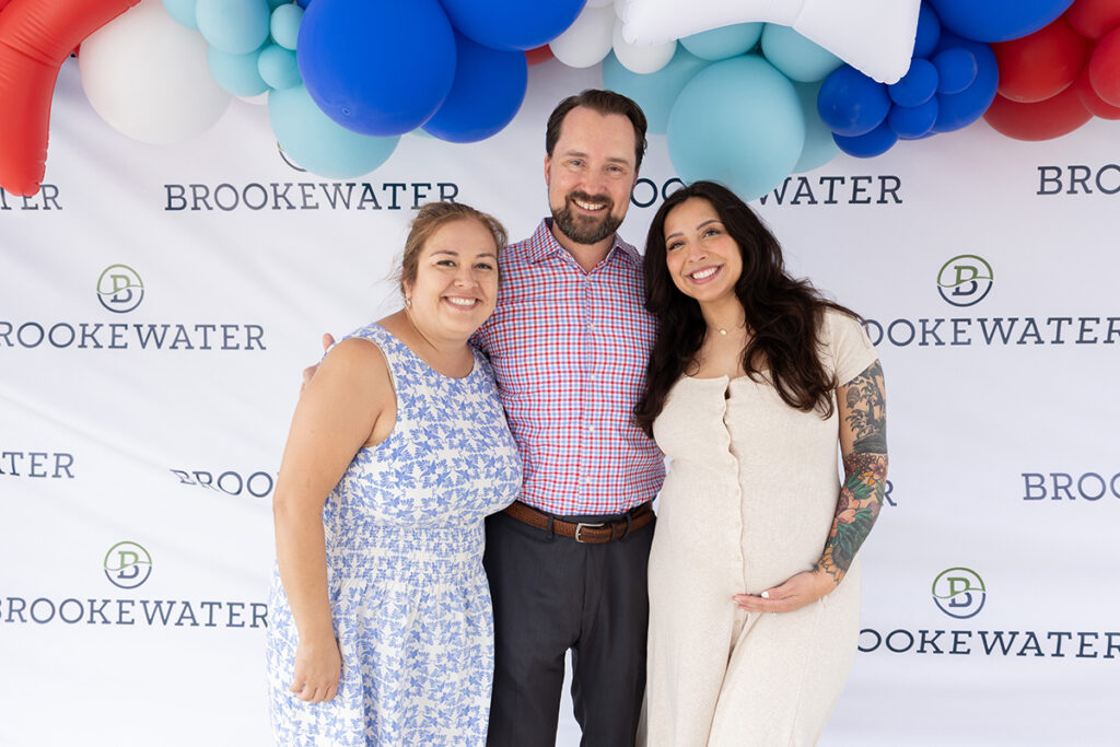 Three adults pose and smile by a BrookeWater backdrop in a master-planned community event.