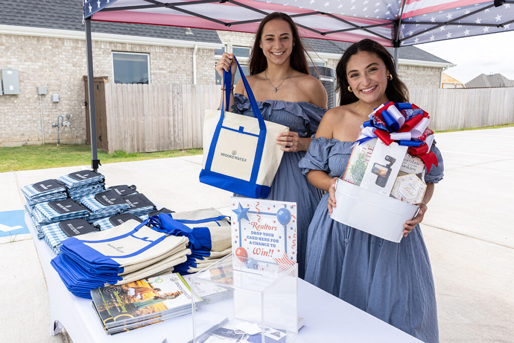 Two women at an outdoor booth for Brookewater display branded tote bags and a raffle gift basket.