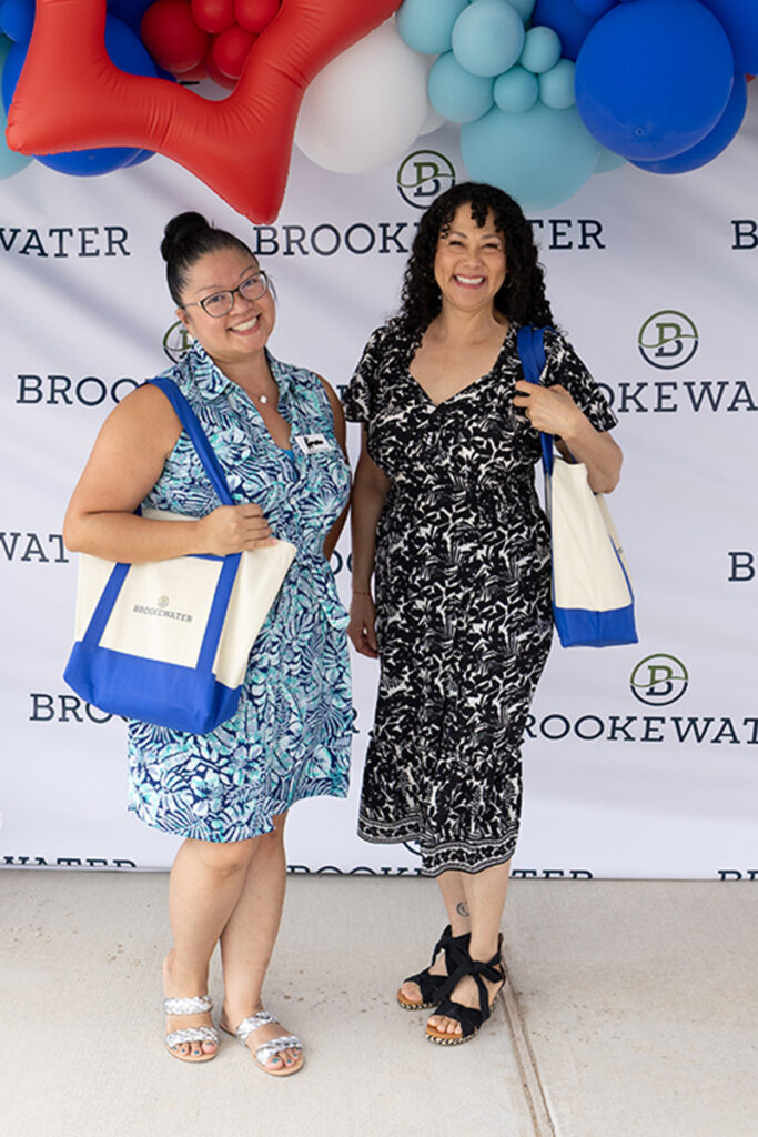Two women with Brookewater totes pose in front of a backdrop near Richmond, TX.