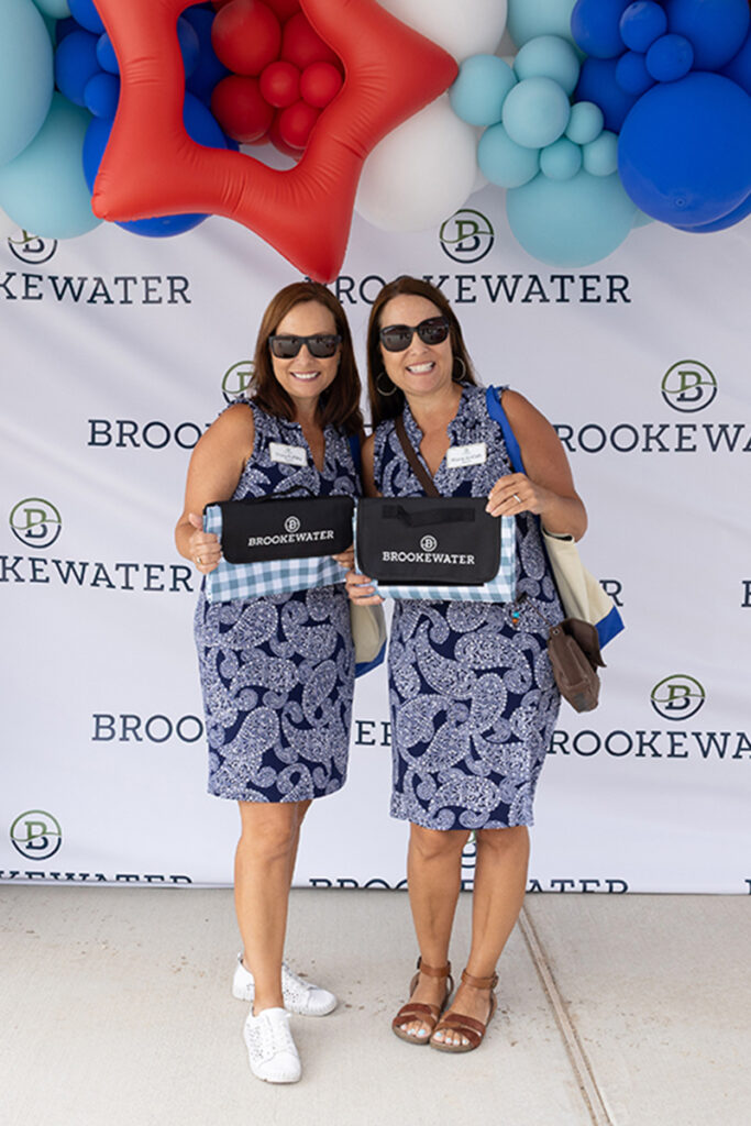 Two women in blue dresses pose with Brookewater bags near Richmond TX and branded balloons.