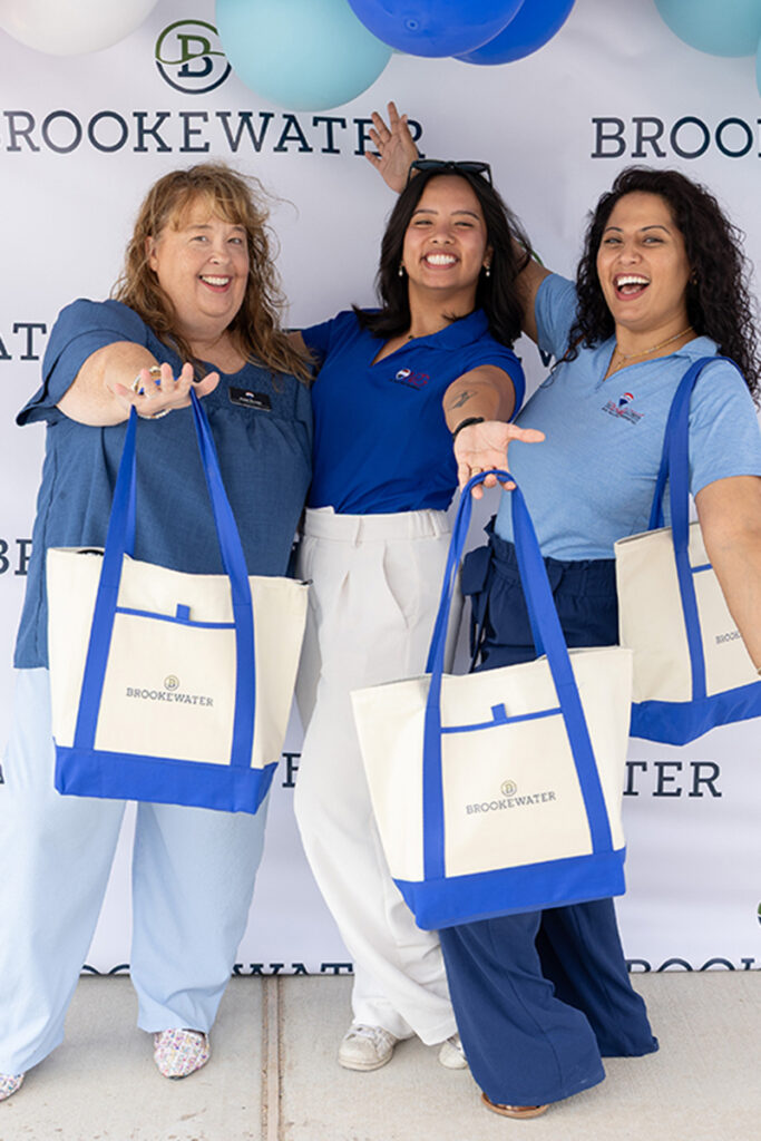 Three women smile with BrookeWater totes near Richmond TX, posing by a branded backdrop.