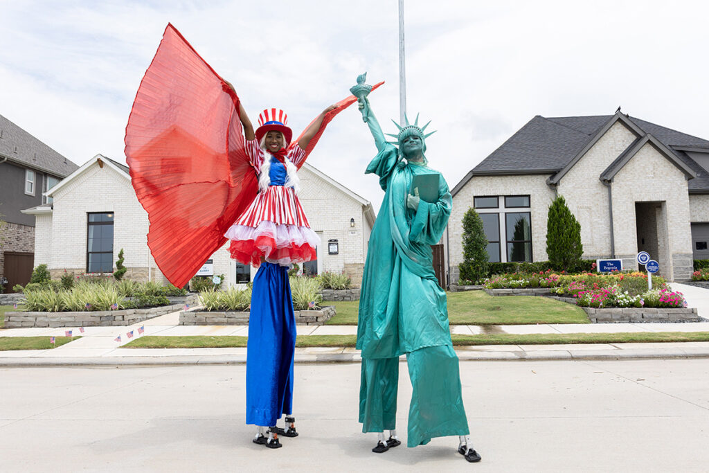 Two people on stilts as Uncle Sam and Lady Liberty stand on a suburban Brookewater street.