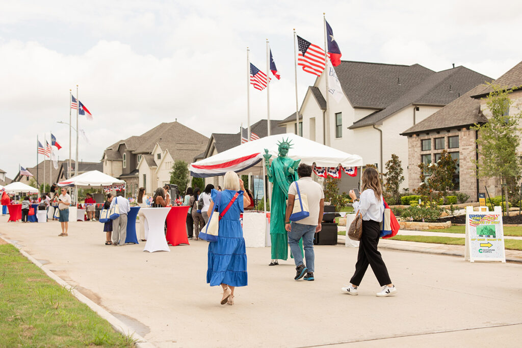 People walk near Richmond TX along a at Brookewater street with tents, flags, and a Statue of Liberty performer.