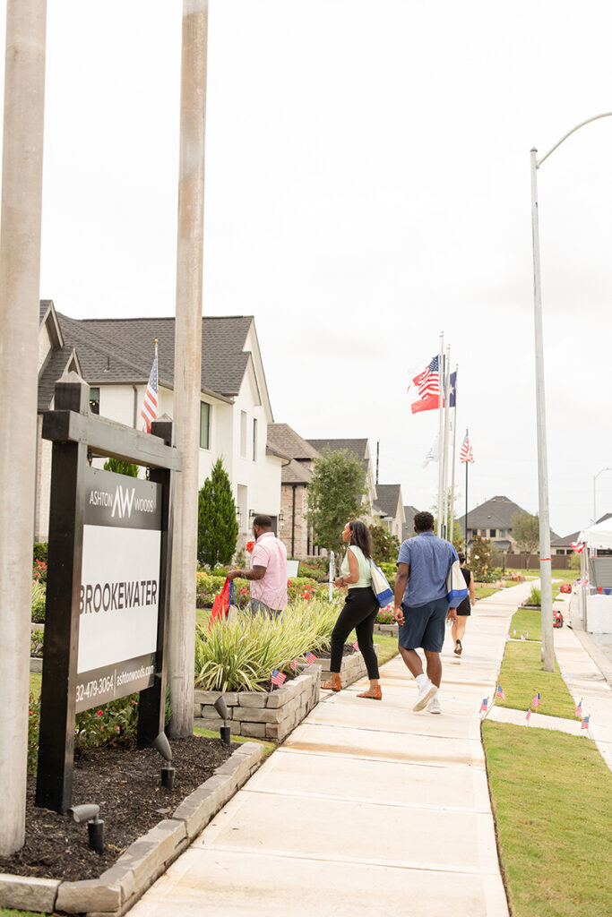 People walk by houses and a BrookeWater master-planned community sign with flags displayed.