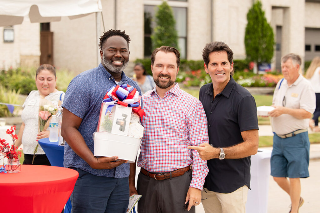 Three men pose outdoors at Brookewater near Richmond TX, one holds a gift basket with a patriotic bow.