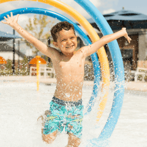 Smiling boy in swim trunks runs through splash pad water jets in Brookewater, near Richmond TX.