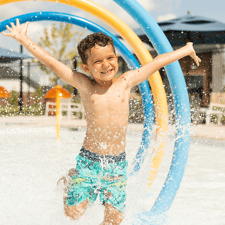 Smiling boy in swim trunks runs through splash pad water jets in Brookewater, near Richmond TX.