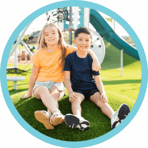 Two children sit on artificial grass at a playground in a vibrant master-planned community.