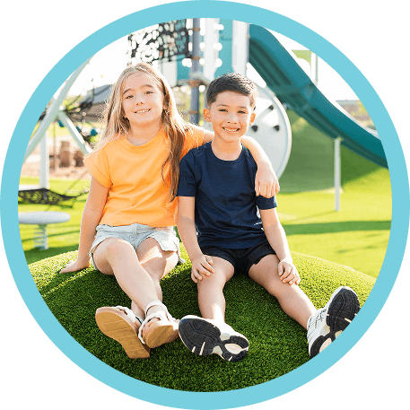 Two children sit on artificial grass at a playground in a vibrant master-planned community.